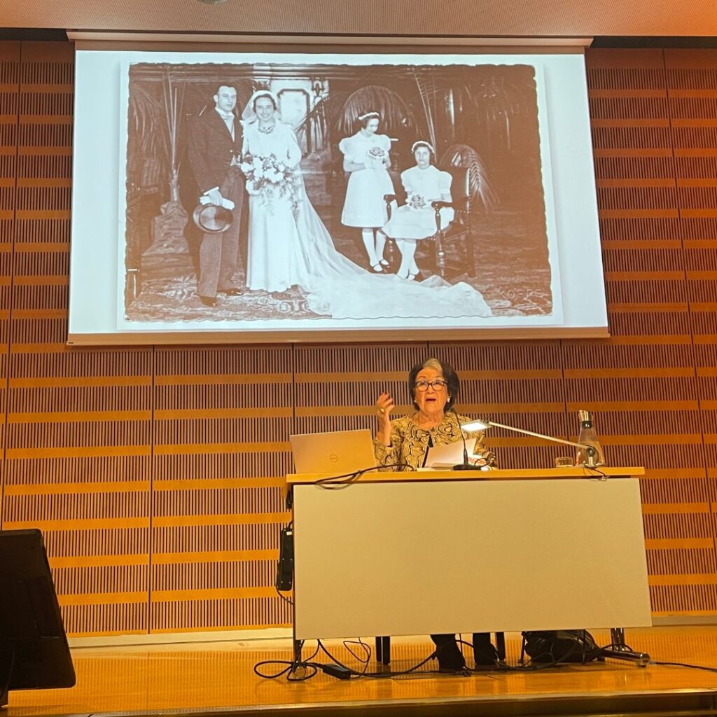 Vortrag in einem Saal: Eine Referentin spricht am Pult, darüber ist ein historisches Foto (Hochzeitsgruppe) groß projiziert.