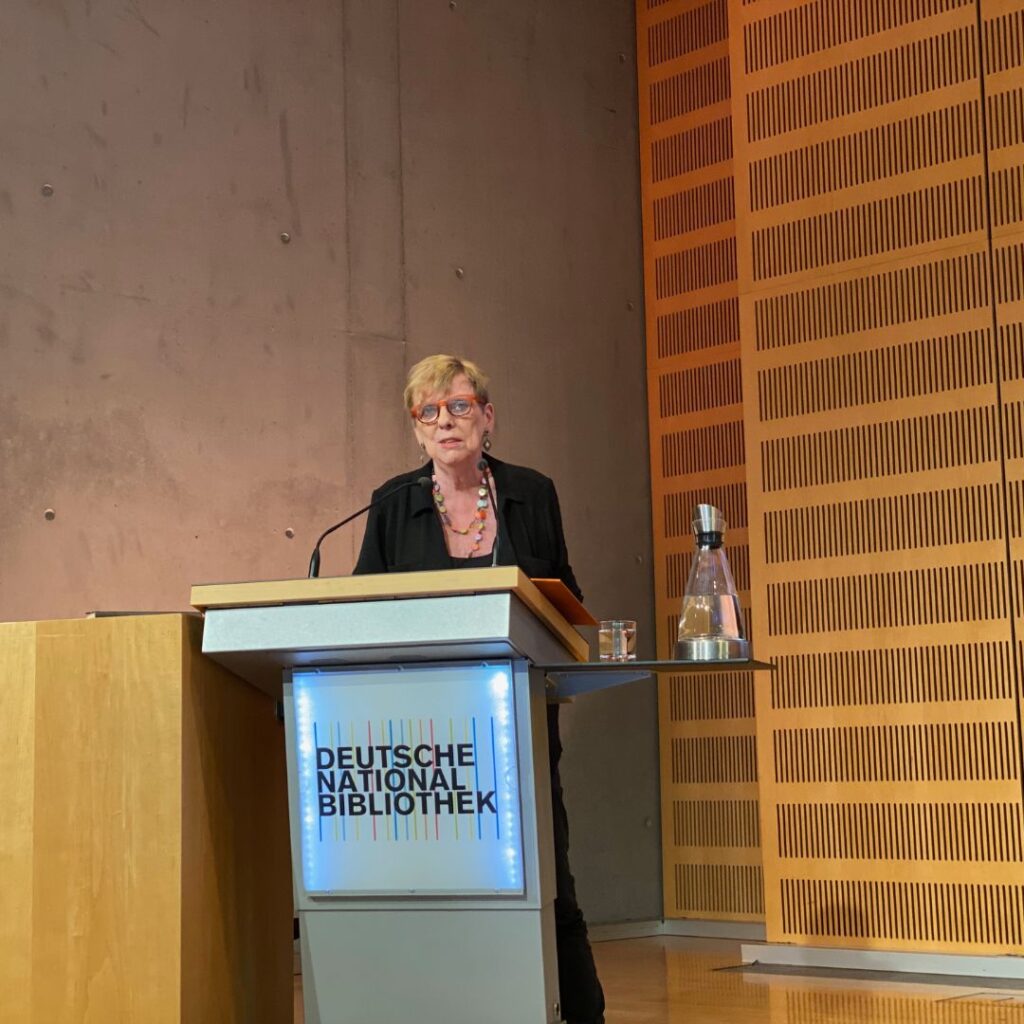 Eine Rednerin steht am Podium mit dem Logo „DEUTSCHE NATIONALBIBLIOTHEK“ und spricht ins Mikrofon; auf dem Podium steht eine Wasserflasche.