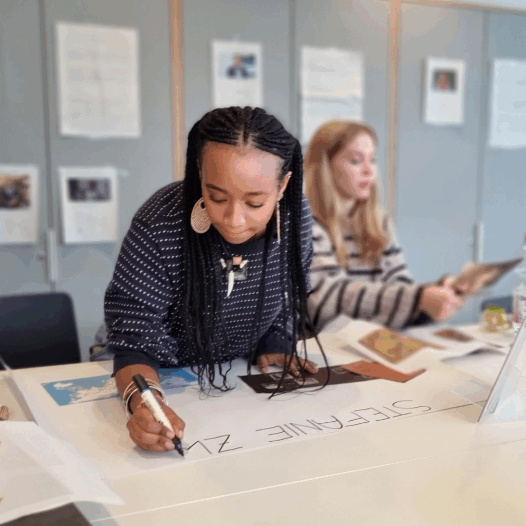 Workshop-Szene: Eine Teilnehmerin beschriftet ein großes Plakat mit „STEFANIE Z“, im Hintergrund sitzt eine weitere Person am Tisch.
