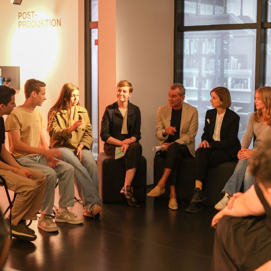 Gesprächsrunde mit mehreren Teilnehmenden in einem Ausstellungsraum; an der Wand steht „POST-PRODUKTION“.