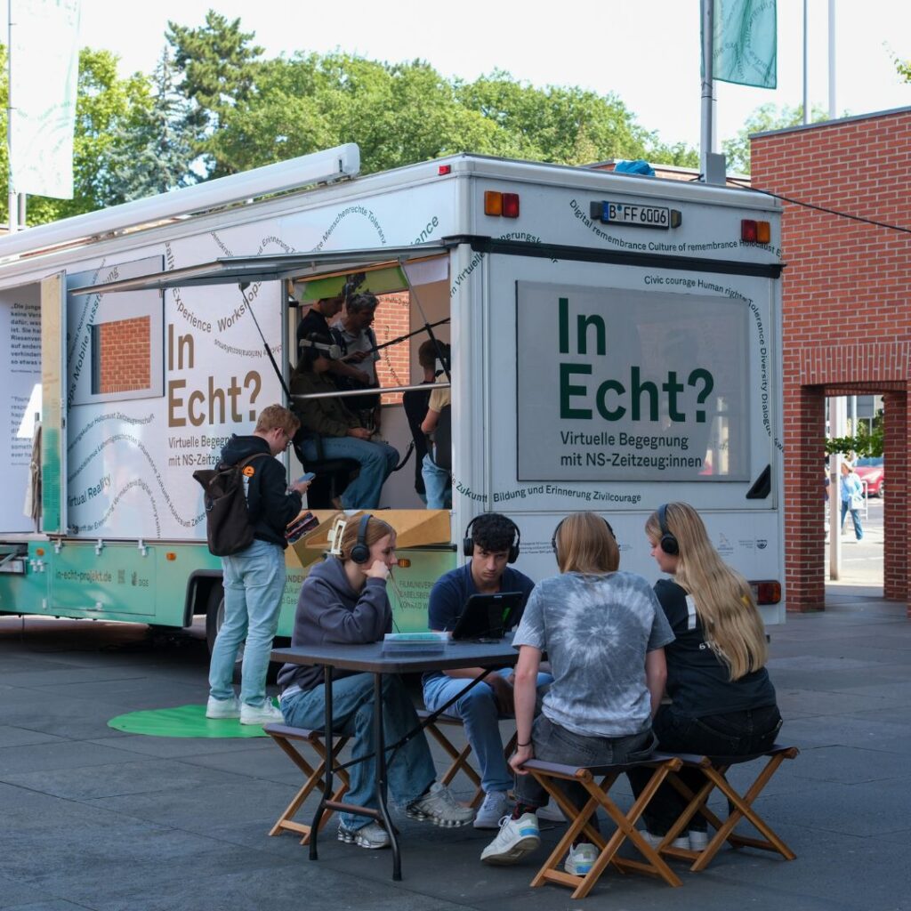 Schülergruppe mit Kopfhörern sitzt an einem Tisch vor einem Info-Truck mit der Aufschrift „In Echt? Virtuelle Begegnung mit NS-Zeitzeug:innen“.