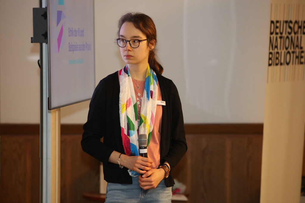 Eine Person mit farbigem Tuch um den Hals hält ein Mikro, links und rechts von ihr stehen ein Bildschirm mit einem unscharfen Logo und ein Roll-Up mit dem Logo der "Deutschen Nationalbibliothek"