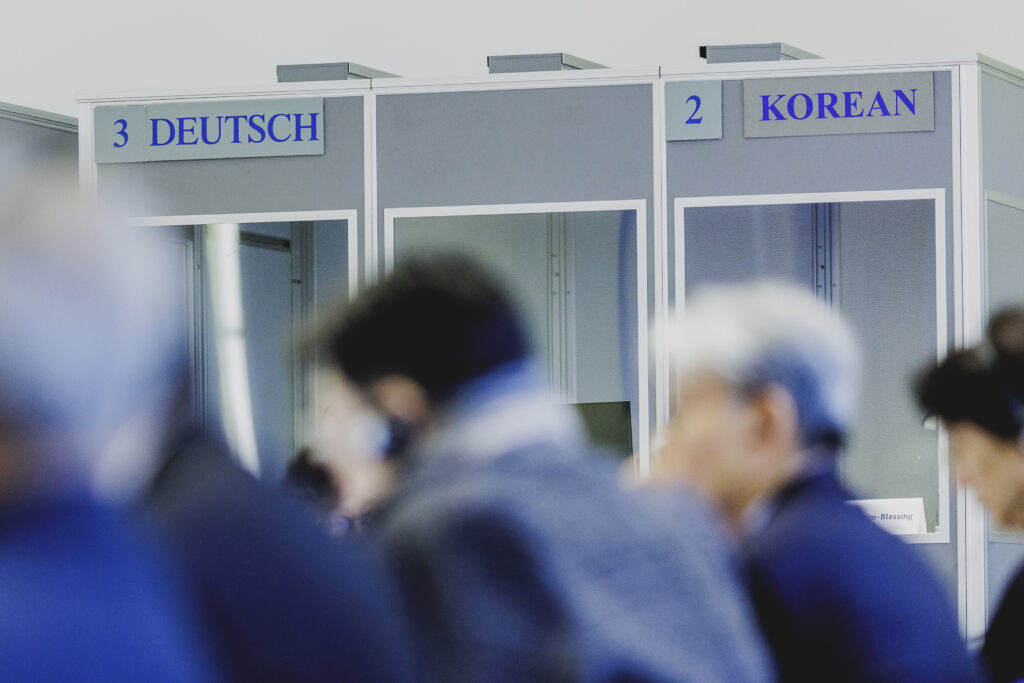 Im Vordergrund sind verschwommen Menschen zu erkennen. Im Hintergrund sieht man deutlich graue Kabinen mit Glasfenstern, die linke ist beschriftet mit "3 Deutsch" und die rechte ist beschriftet mit "2 Korean".