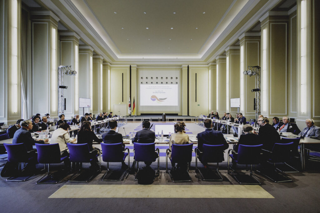 In einem großen, hellen Saal mit vielen Säulen sitzen viele Menschen an einem großen U-förmigen Tisch und reden miteinander.