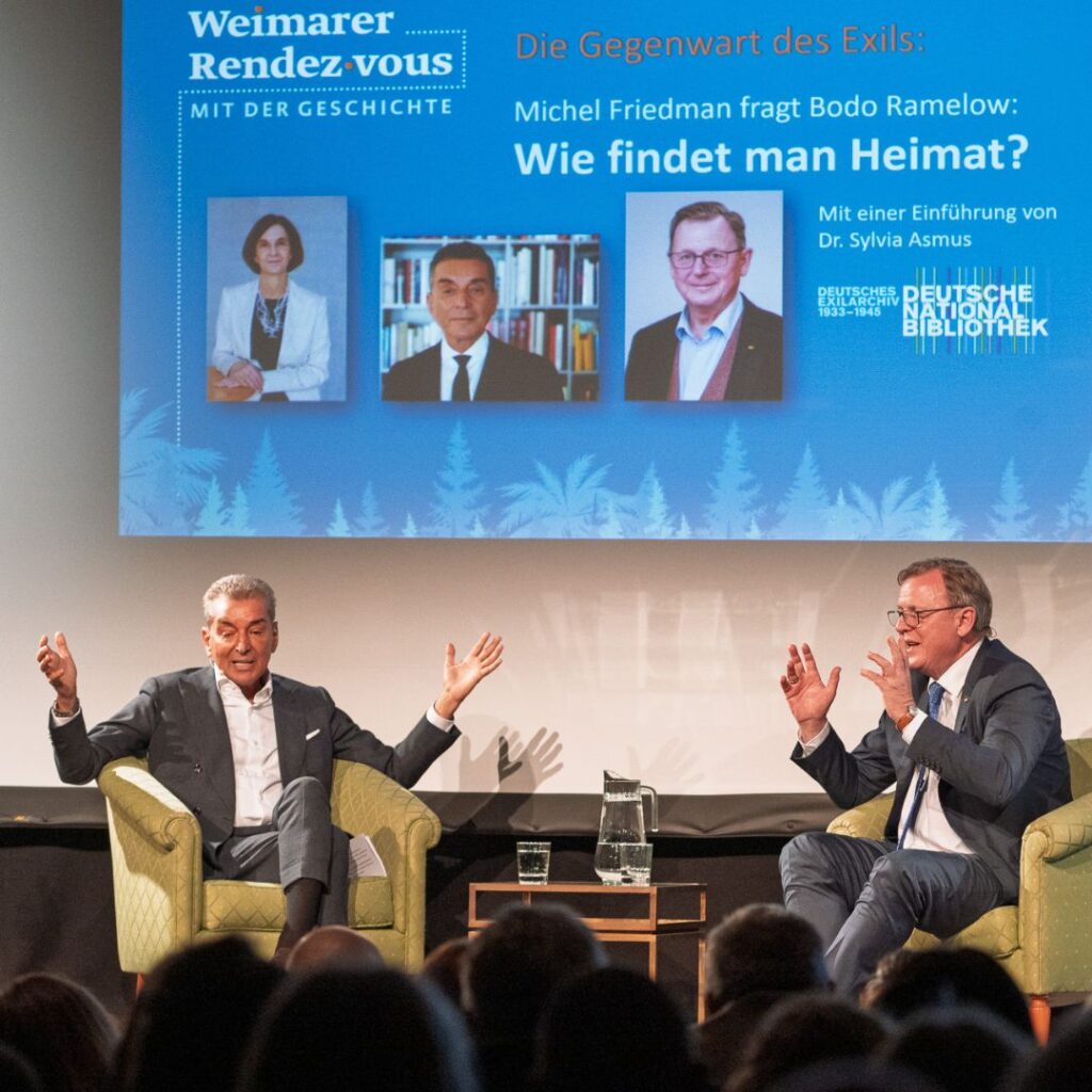 Zwei Männer sitzen in Sesseln auf einer Bühne im Gespräch, darüber eine Veranstaltungsfolie mit Titel und Porträtfotos.