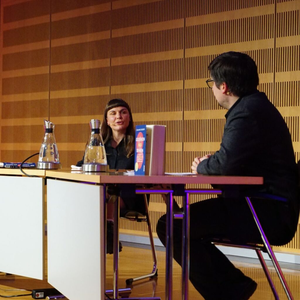 Zwei Personen im Gespräch an einem Tisch auf der Bühne, mit Mikrofon, Wasserflaschen und einem aufgestellten Buch.