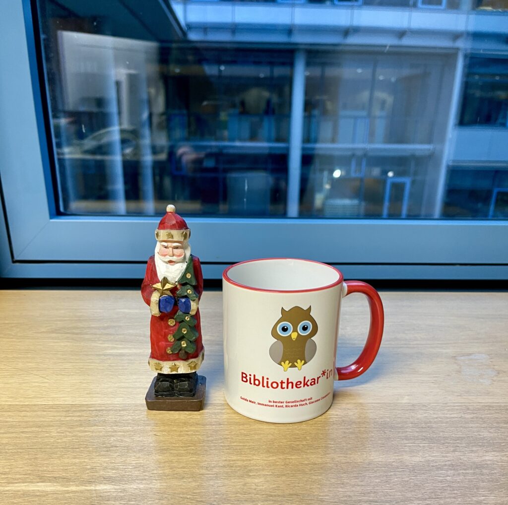 Ein Weihnachtsmann aus Holz und eine Tasse mit der Aufschrift Bibliothekar*in und eine Eule