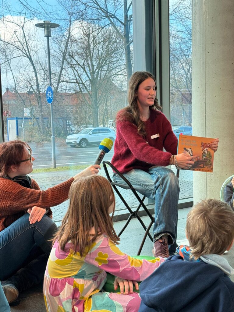 Eine Person zeigt einem Publikum das Cover eines Buches, neben ihr sitzt eine Person, die ein Mikro in ihre Richtung hält