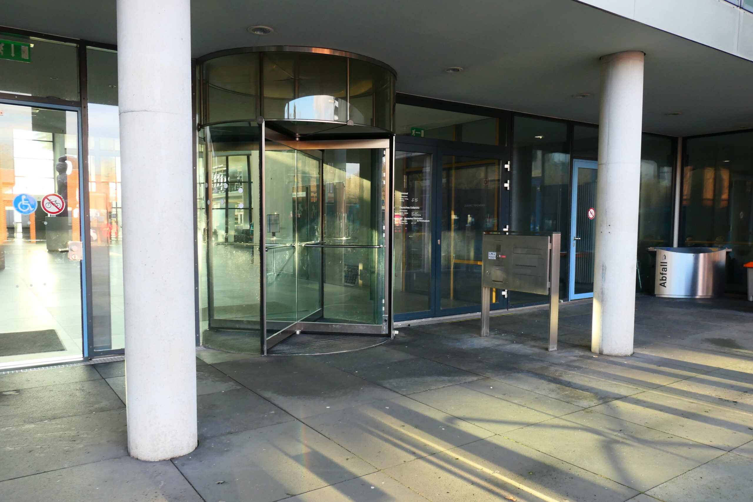Man sieht eine Drehtür aus Glas. Die Wände rechts und links davon bestehen ebenfalls aus einer großen Fensterfront. Rechts und links neben der Drehtür stehen zwei weiße Beton-Säulen, die die Überdachung des Eingangsbereiches tragen.