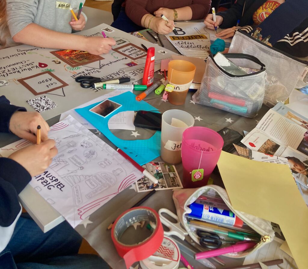 Hände arbeiten an einem Tisch mit Plakaten, Stiften, KLleber, Tape, Postkarten. Auch Getränkebecher sind zu sehen.