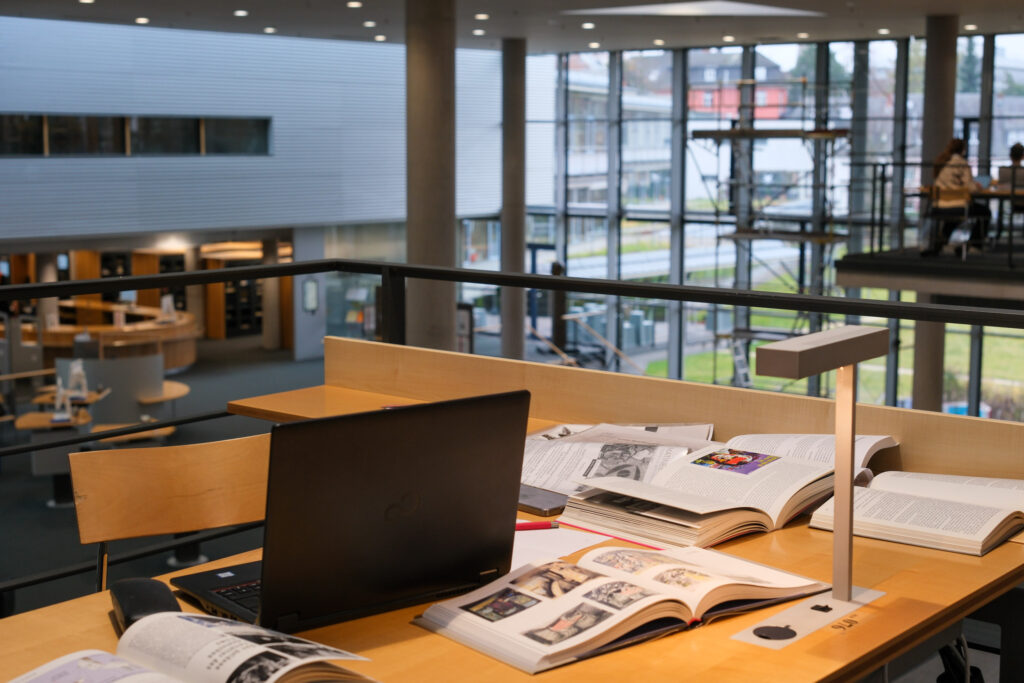 Man sieht von der Empore aus auf den Lesesaal der Deutschen Nationalbibliothek in Frankfurt. Im Vordergrund steht ein Schreibtisch, an dessen vorderer Kante eine Leselampe befestigt ist. In der Mitte des Schreibtisches steht ein aufgeklapptes Notebook. Um das Notebook herum sind verschiedene, aufgeschlagene Bücher verteilt. Der Stuhl hinter dem Schreibtisch ist leer. 
