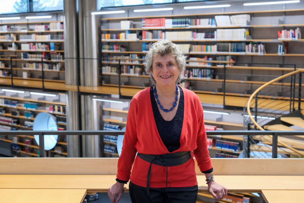 Man sieht eine Frau, die in die Kamera lächelt. Sie trägt eine schwarze Bluse mit einer roten Strickjacke und eine lange Kette aus großen blau-grünen Steinen. Die Frau steht an ein Geländer gelehnt. Hinter ihr sieht man eine über zwei Etagen gehende Bücherwand