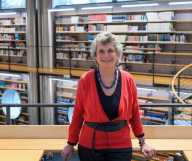 Man sieht eine Frau, die in die Kamera lächelt. Sie trägt eine schwarze Bluse mit einer roten Strickjacke und eine lange Kette aus großen blau-grünen Steinen. Die Frau steht an ein Geländer gelehnt. Hinter ihr sieht man eine über zwei Etagen gehende Bücherwand
