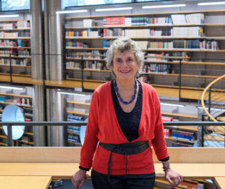 Man sieht eine Frau, die in die Kamera lächelt. Sie trägt eine schwarze Bluse mit einer roten Strickjacke und eine lange Kette aus großen blau-grünen Steinen. Die Frau steht an ein Geländer gelehnt. Hinter ihr sieht man eine über zwei Etagen gehende Bücherwand