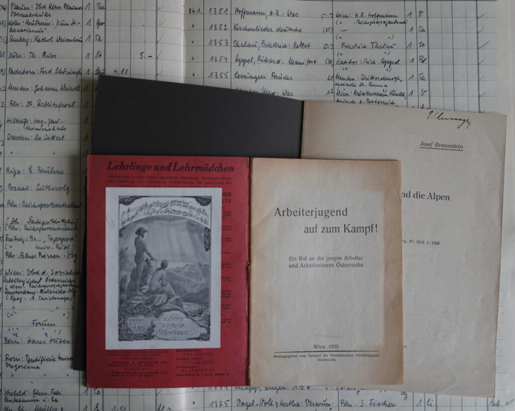 Auf einem aufgeschlagenen Zugangsbuch der Deutschen Bücherei liegen zwei aufgeschlagene Bücher. Im einem davon klebt im Vorsatz das Exlibris von Wilhelm Ellenbogen, im zweiten ist auf dem Titelblatt das Autogramm von Ellenbogen erkennbar.