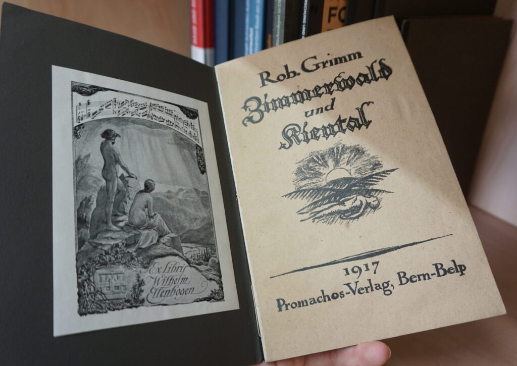 Eine Hand hält der Kamera ein aufgeschlagenes Buch entgegen, in dessen Vorsatz das Exlibris von Wilhelm Ellenbogen klebt. Im Hintergrund ist ein Bücherregal mit mehreren Büchern zur Provenienzforschung erkennbar.