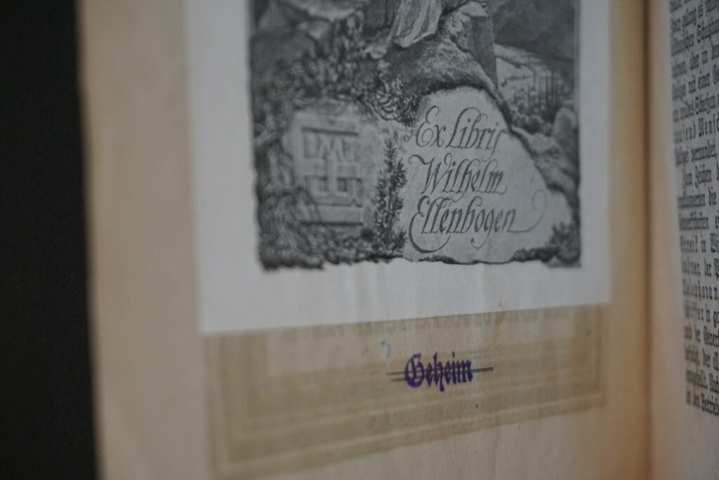Zu sehen ist ein Ausschnitt von Wilhelm Ellenbogens Exlibris, Darunter ist in Frakturschrift das Wort "Geheim" eingestempelt und später mit Bleistift durchgestrichen worden.