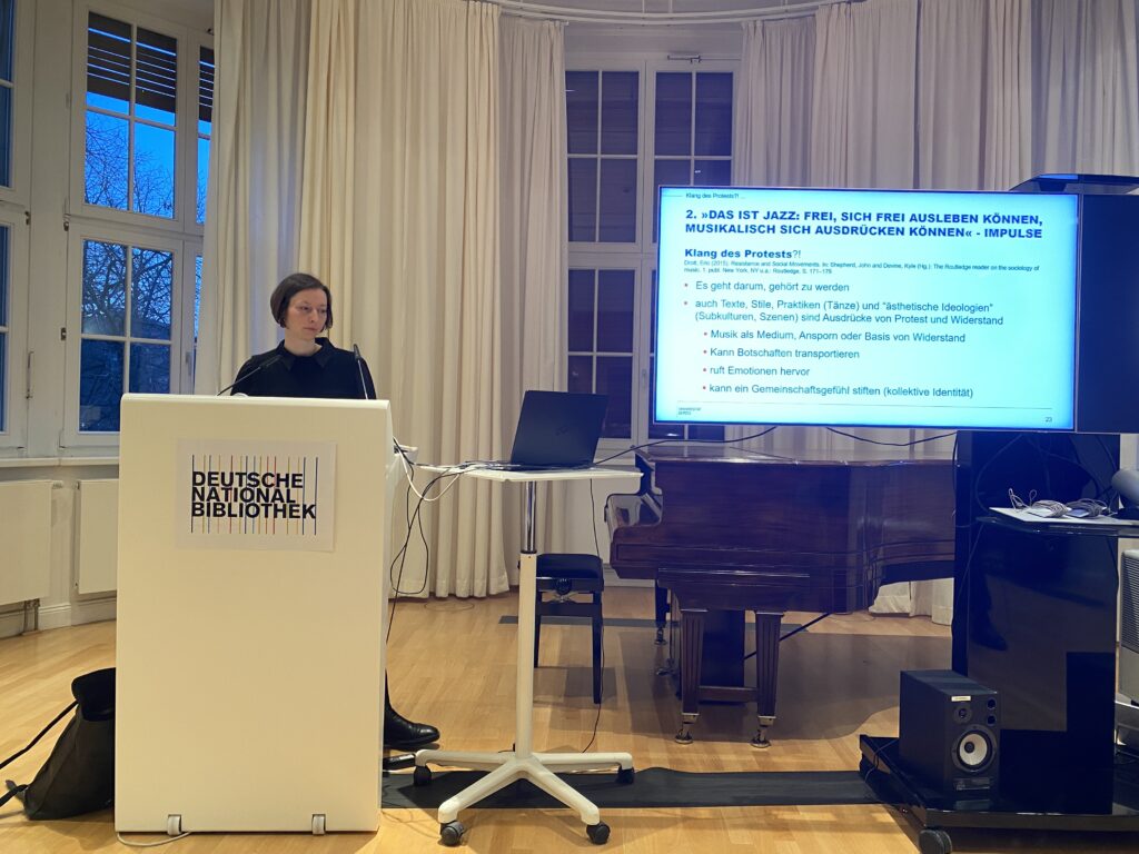 Eine Frau steht am Pult und schaut auf den Laptop. Die Präsentation zeigt eine Folie über Jazz als Klang des Protests
