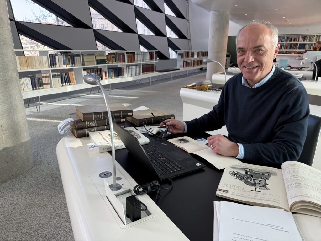 Man sieht einen Mann, der den Betrachter anlächelt. In der rechten Hand hält er eine Brille. Er sitzt an einem Schreibtisch in modernem Design. Vor ihm steht ein aufgeklapptes Notebook und verschiedene aufgeschlagene Texte. Eine der vergilbten Seiten zeigt die Zeichnung einer Maschine. Auf der rechten Seite des Tisches liegt ein Stapel alt aussehender Bücher mit braunen Einbänden und goldenen Verzierungen. Vor dem Mann an der Tischkante ist eine chromfarbene Leselampe befestigt. Im Hintergrund sieht man eine Fensterfront mir eine Reihe halbhoher Bücherregale davor. Die Fenster sind dreieckig. Wobei immer ein gleich großes Rechteck diagonal geteilt und jeweils in der unteren Hälfte verglast wurde. 
