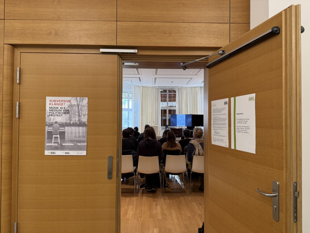 Blick von außen in den Vortragsraum, die Stuhlreihen sind gefüllt; an der Tür hängt das Plakat der Tagung.