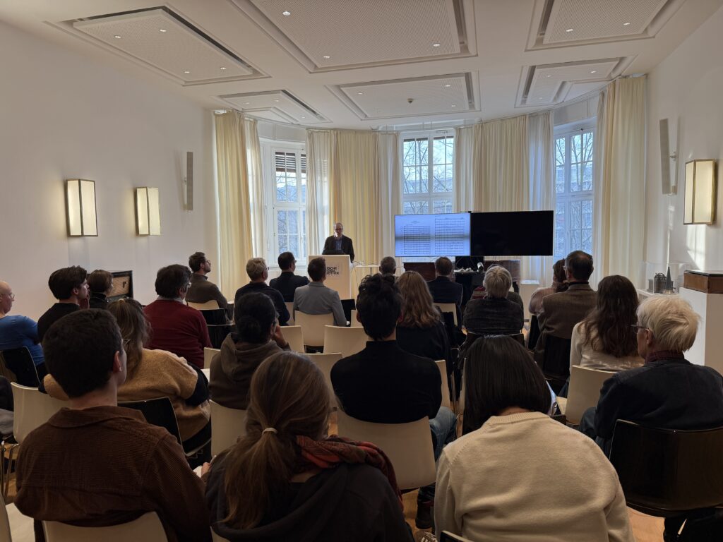 Ein Referent steht am Rednerpult, der Fokus des Bildes liegt auf dem Publikum des gefüllten Raums.