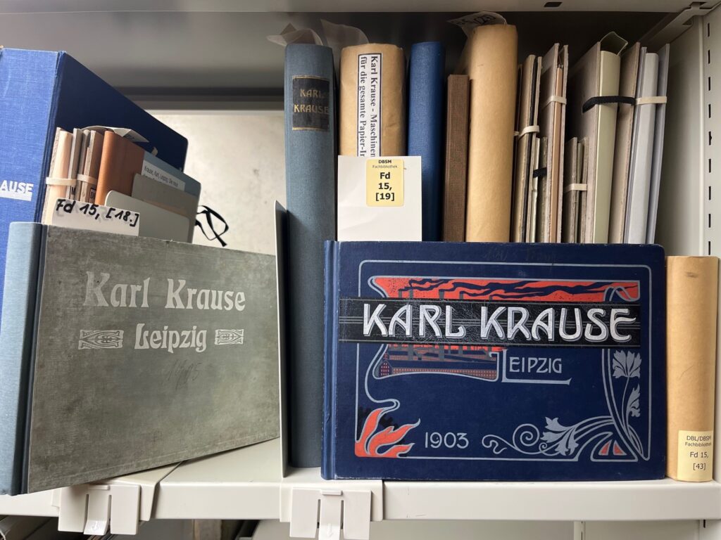 Man sieht ein Regal im Magazin der Deutschen Nationalbibliothek Leipzig. Die beiden Bücher im Vordergrund tragen den Titel "Karl Krause Leipzig" Das rechte Exemplar dazu noch die Jahreszahl 1903. Hinter diesen beiden Büchern stehen weitere alt wirkende Publikationen, die ebenfalls mit dem Namen "Karl Krause" betitelt sind.