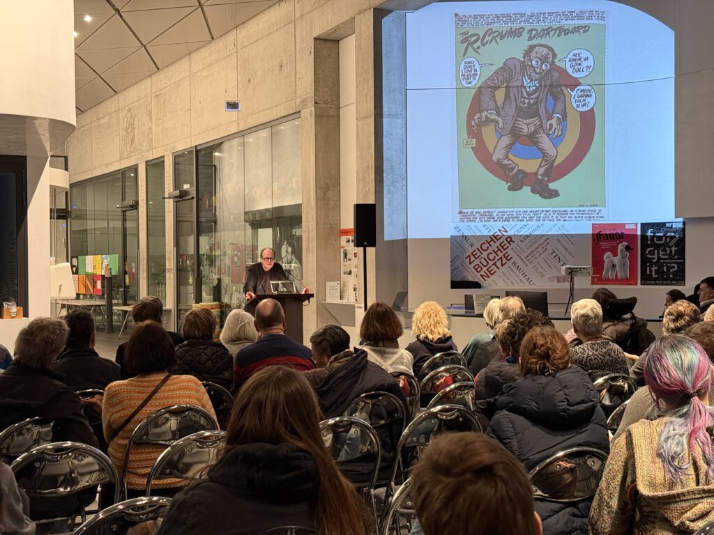 Zuschauer sitzen in Reihen. Im Hintergrund steht ein Mann am Pult, der einen Vortrag hält. Auf der Wand ist ein Selbstporträt von Crumb projiziert.