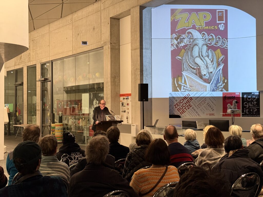 Zuschauer sitzen in Reihen. Im Hintergrund steht ein Mann am Pult, der einen Vortrag hält. Auf der Wand ist das Titelblatt von Zap-Comib Nummer 6 projiziert.