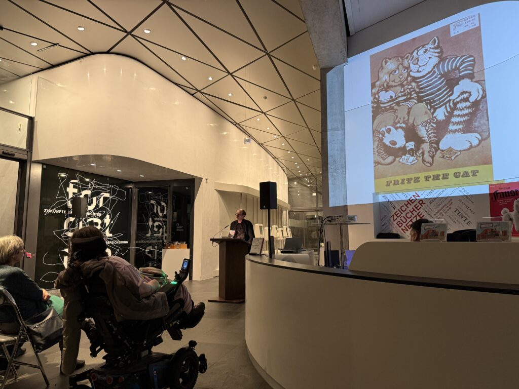 Ein moderes Foyer mit Zuschauern und einem Rednern. An der Wand ist ein Comic mit zwei Katzen pojiziert. Darunter steht "Fritz the Cat".