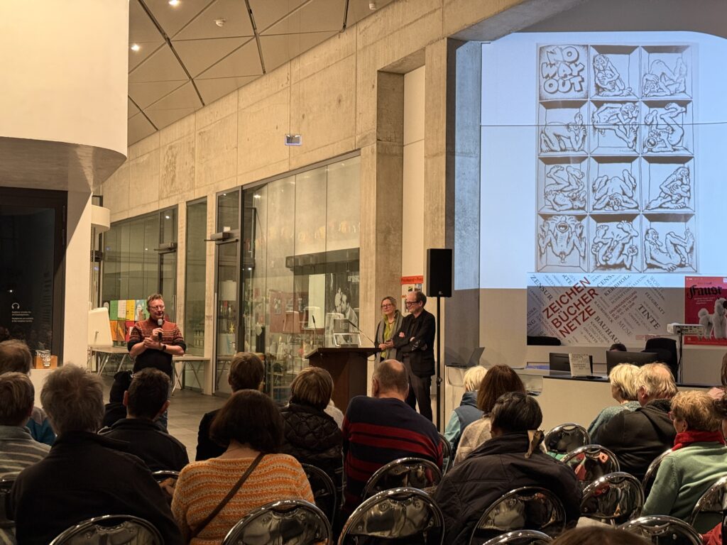 Zuschauer sitzen in Reihen. Im Hintergrund stehen drei Personen, die miteinander reden. Auf der Wand ist ein Comic von Crumb projiziert.