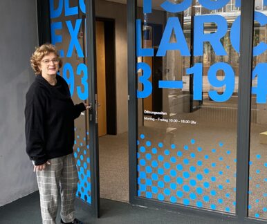 Eine Frau lächelt in die Kamera. Sie schaut den Betrachter an, während sie eine Glastür mit Aufdruck öffnet. Auf der Tür steht in blauen Buchstaben "Deutsches Exilarchiv 1933 - 1945".