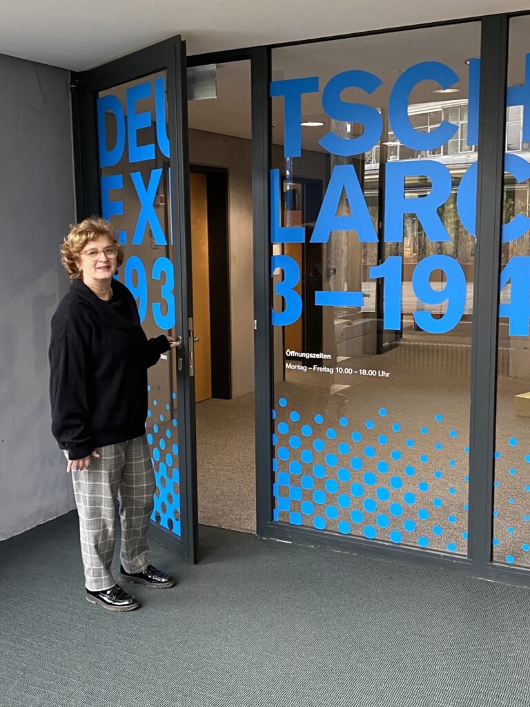 Eine Frau lächelt in die Kamera. Sie schaut den Betrachter an, während sie eine Glastür mit Aufdruck öffnet. Auf der Tür steht in blauen Buchstaben "Deutsches Exilarchiv 1933 - 1945".