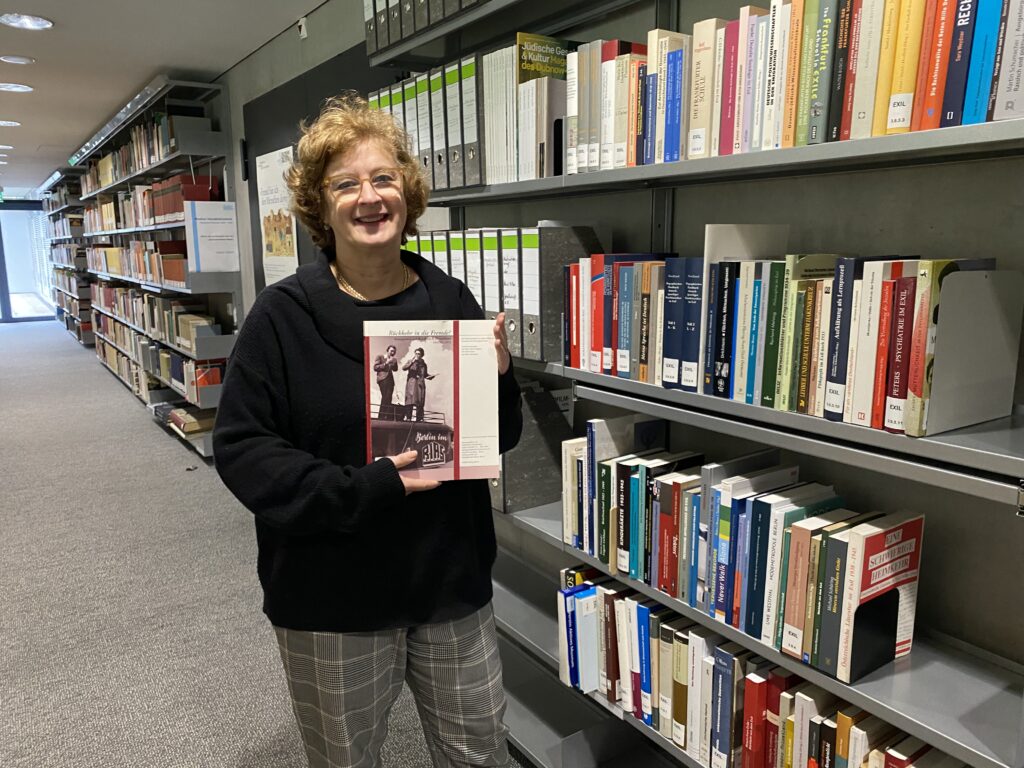 Eine Frau steht vor der Handbibliothek des Deutschen Exilarchives der deutschen Nationalbibliothek in Frankfurt am Main. Sie lächelt in die Kamera und präsentiert dem Betrachter ein Buch. In den Regalen, die sich in der rechten Bildhälfte in den Hintergrund fortsetzen stehen verschiedene Bücher zu Exilthemen.