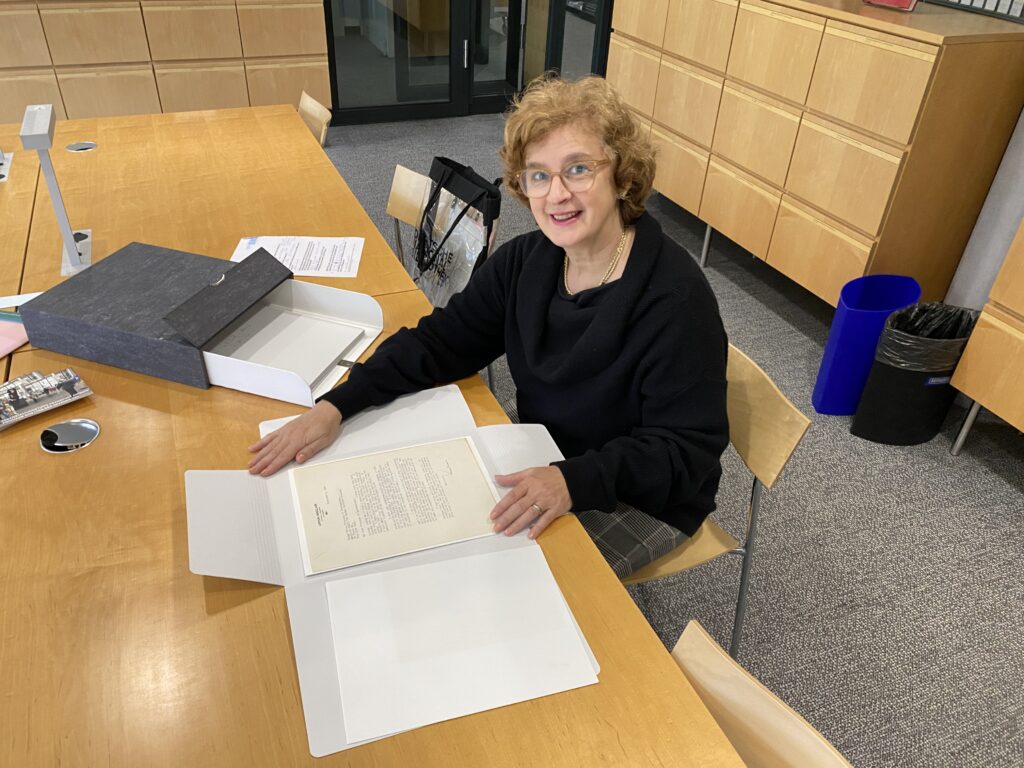 Eine Frau sitzt an einem Schreibtisch und lächelt in die Kamera. Vor ihr auf dem Tisch steht ein geöffneter Archivkarton mit mehreren Archivmappen darin. Eine der Mappen liegt geöffnet vor der Frau auf dem Tisch. Auf dem leeren Stuhl neben ihr steht eine durchsichtige Tasche aus robustem Plastik, die das Logo der Deutschen Nationalbibliothek trägt.