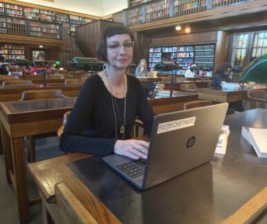 Eine Frau sitzt an einem Schreibtisch aus Holz. Vor ihr steht ein aufgeklapptes Notebook, um das mehrere aufgeschlagene Bücher verteilt sind. Im Hintergrund sieht man einen großen Raum, in dem weitere Schreibtische hintereinander aufgereiht stehen. Hier sitzen weitere Leute und lesen oder arbeiten. Die rückwärtige Wand besteht aus einem großen Bücherregal mit vielen alt aussehende Medienwerken. In der Rechten Bildhälfte befindet sich eine verglaste Eingangstür. Auch hier ist die Architektur älter.