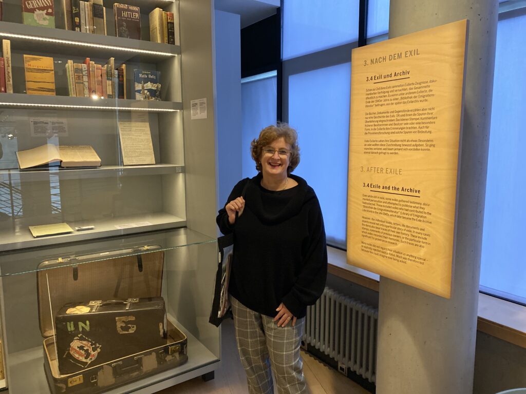 Eine Frau steht in den Ausstellungsräumen des Deutschen Exilarchivs in Frankfurt. Sie lächelt in die Kamera und trägt eine der Bibliothekstaschen mit dem Logo der DNB über der Schulter. Auf der rechten Seite des Bildes befindet sich eine Informationstafel mit der Überschrift „Nach dem Exil“. In der linken Bildhälfte sieht man eine hohe Vitrine. Sie enthält einen alten Reisekoffer, ein maschinengeschriebenes Manuskript , ein aufgeschlagenes Buch so wie weitere Bücher, die Rücken an Rücken auf einem Regalbrett aufgereiht sind.