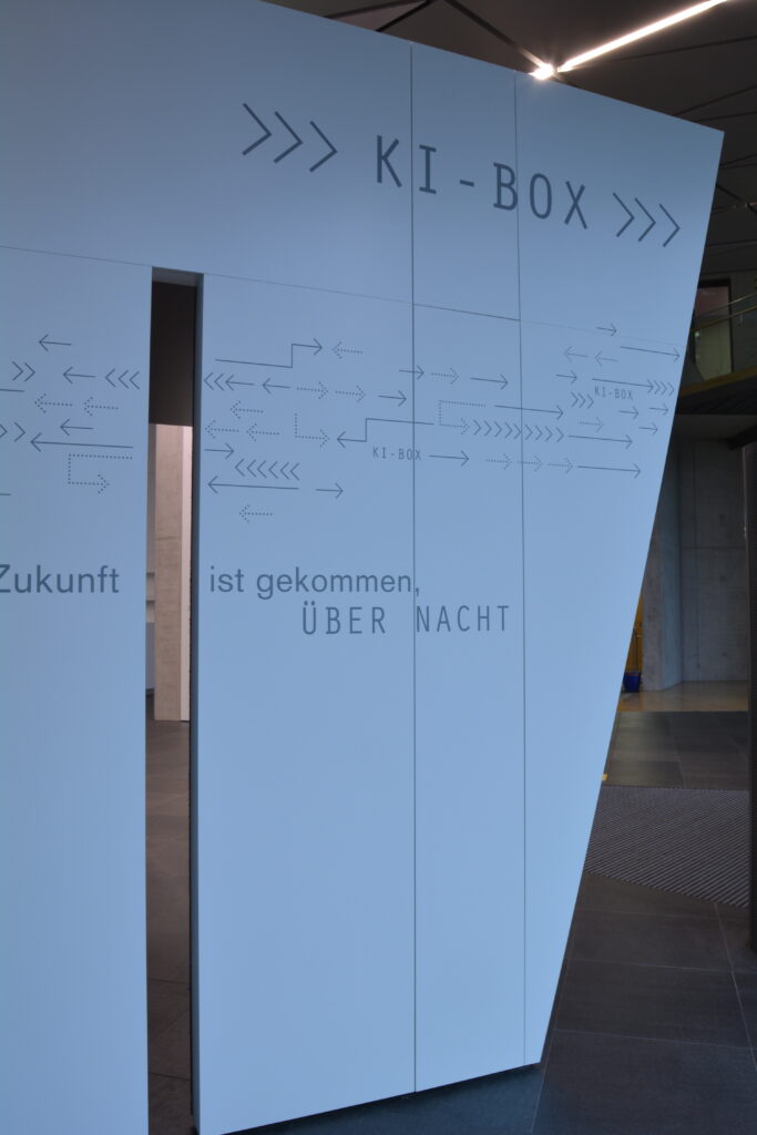 Eine weiße Box mit einem schmalen Spalt in einer Seite und der Aufschrift "KI-Box" steht in einem indirekt beleuchteten, großen Raum.