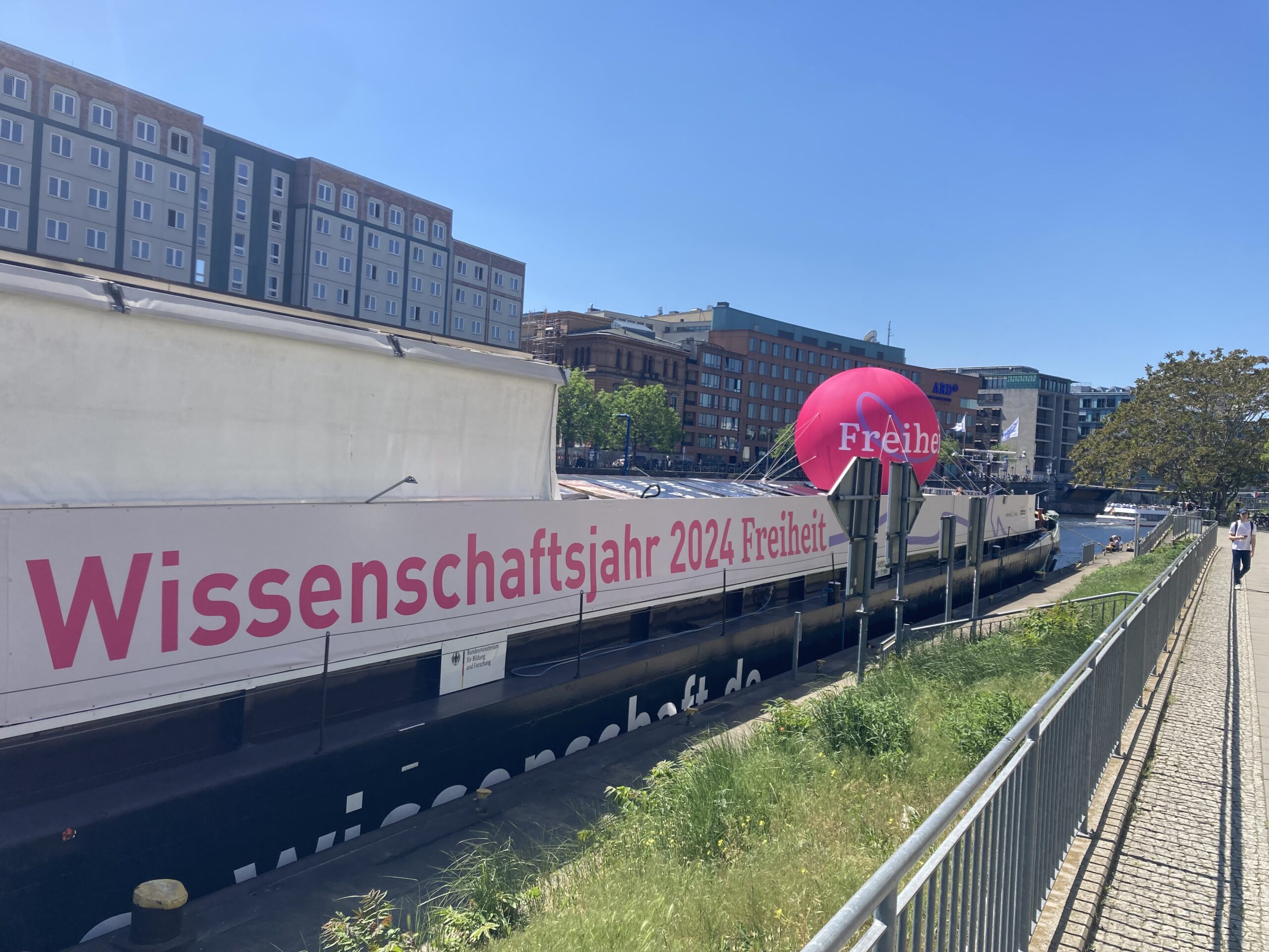 Ein Binnenschifffahrtsschiff an einer Anlegestelle. Auf der Seite ist ein großes Banner angebracht auf dem in pinker Schrift steht: "Wissenschaftsjahr 2024 Freiheit". Weiterhin ist ein großer pinker Ballon mit der Aufschrift "Freiheit" auf dem Schiff angebracht.