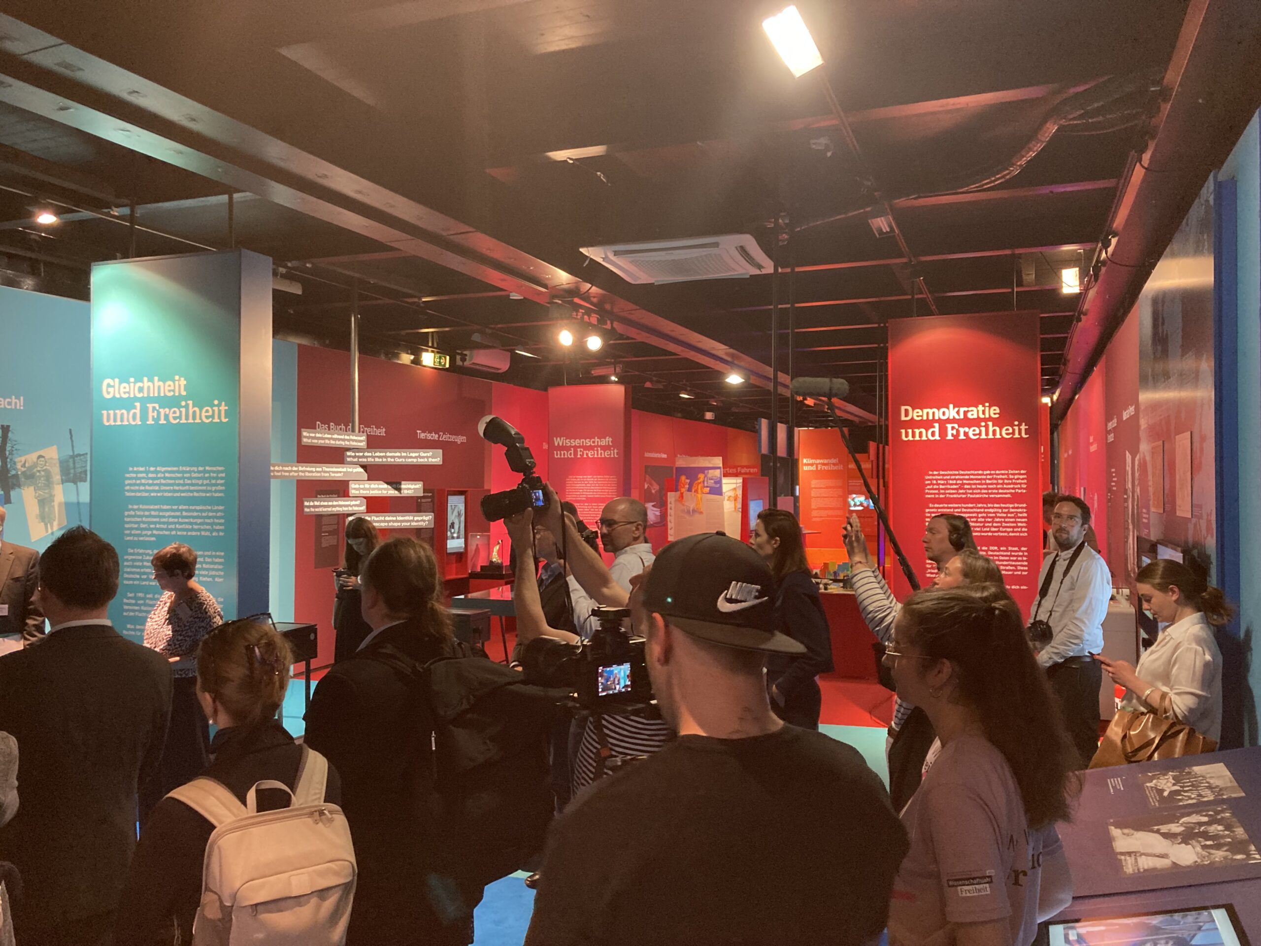 Menschen mit Fotoapparaten und Mikrophonen stehen dicht an dich in einem Ausstellungsraum. Im Hintergrund erkennt man Themenüberschriften, wie "Gleichheit und Freiheit" sowie "Demokratie und Freiheit".