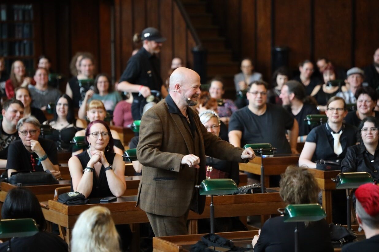 Viele Menschen sitzen an hölzernen Tischen und hören einem Mann zu, der mittig steht und redet.