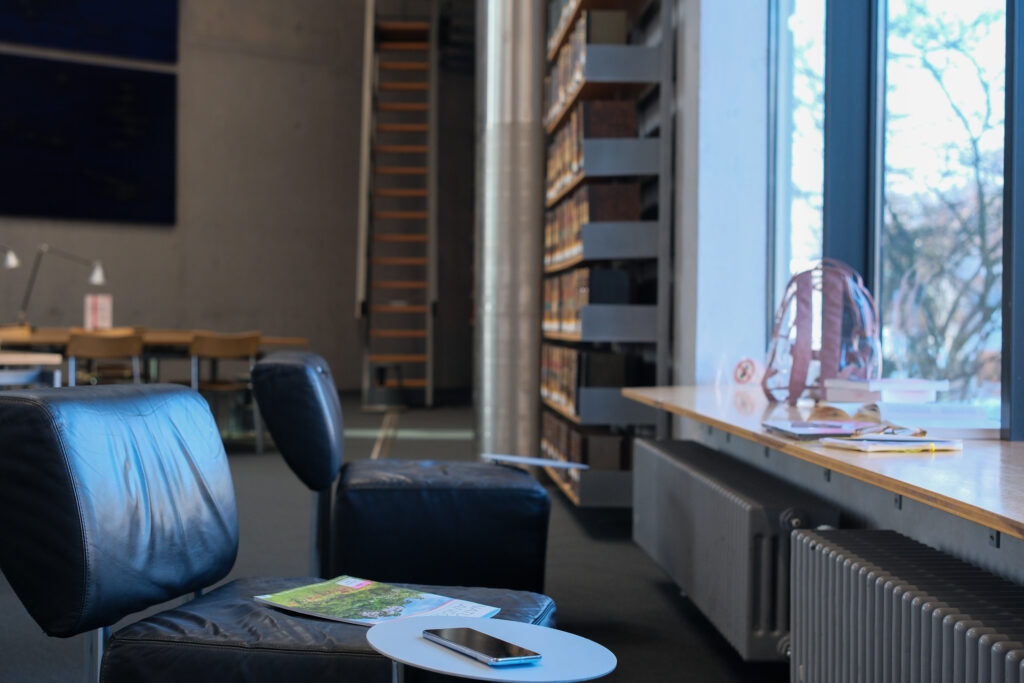 Man sieht den Lesesaal der Deutschen Nationalbibliothek in Frankfurt am Main. Auf der rechten Seite des Bildes befindet sich ein großes Fenster mit einer tiefen Fensterbank. Unter der Fensterbank sind zwei graue Heizkörper montiert. Dem Fenster gegenüber stehen zwei schwarze Polsterstühle, an d3nen ein kleiner, runder Tisch angebracht ist. Beide Plätze sind leer, aber auf dem vorderen liegen eine Zeitrischt und ein Smartphone. Auf der Rechten Seite im Hintergrund sieht man ein deckenhohes Regal voller alt aussehender Bücher.
