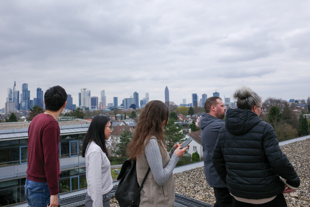 Im Vordergrund steht eine Gruppe von fünf Personen unterschiedlicher Geschlechterzusammensetzung. Eine Frau schaut auf ihr Handy. Im Hintergrund sieht man die Skyline der Stadt Frankfurt am Main.