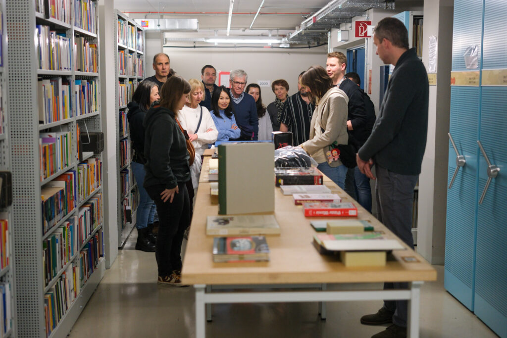 Im Hintergrund steht eine Gruppe von Menschen eng zusammen um das Ende eines langen Tisches herum. Auf diesem Tisch liegen nebeneinander mehrere verschiedene Bücher. Rechts und links wird das Bild von blauen Magazinregalen eingefasst. Links kann man die darin aufgestellten Bücher sehen, rechts die Drehkurbeln, mit denen sie verschoben werden können.
