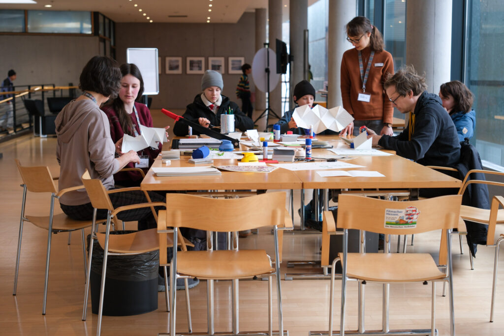 Eine Gruppe von Kindern sitzt an einem großen Tisch. In der Tischmitte liegen verschiedene Bastelutensilien, wie Kleber, Scheren und verschiedene Papiere. Neben einem der Kinder steht eine junge Frau, die ihm etwas erklärt. Eine weitere Sitzt neben einem Kind auf der anderen Seite des Tisches.
