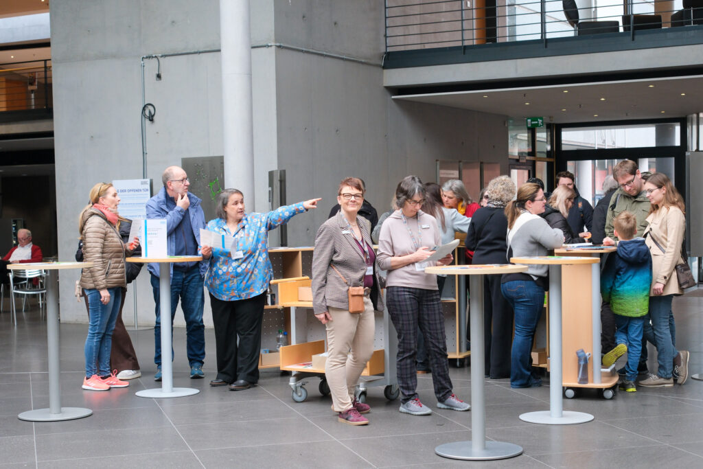 Eine große Gruppe von Menschen steht im Eingangsbereich der Deutschen Nationalbibliothek in Frankfurt am Main. Sie gruppieren sich um mehrere runde Stehtische, die in der hinteren Bildhälfte verteilt sind. In der linken Bildhälfte weist eine Frau in eine Richtung und erklärt einer Personengruppe Etwas.  Unter den Personen sind auch mehrere Kinder.