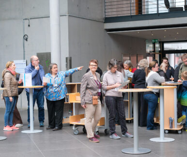 Eine große Gruppe von Menschen steht im Eingangsbereich der Deutschen Nationalbibliothek in Frankfurt am Main. Sie gruppieren sich um mehrere runde Stehtische, die in der hinteren Bildhälfte verteilt sind. In der linken Bildhälfte weist eine Frau in eine Richtung und erklärt einer Personengruppe Etwas.