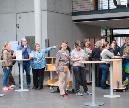 Eine große Gruppe von Menschen steht im Eingangsbereich der Deutschen Nationalbibliothek in Frankfurt am Main. Sie gruppieren sich um mehrere runde Stehtische, die in der hinteren Bildhälfte verteilt sind. In der linken Bildhälfte weist eine Frau in eine Richtung und erklärt einer Personengruppe Etwas.