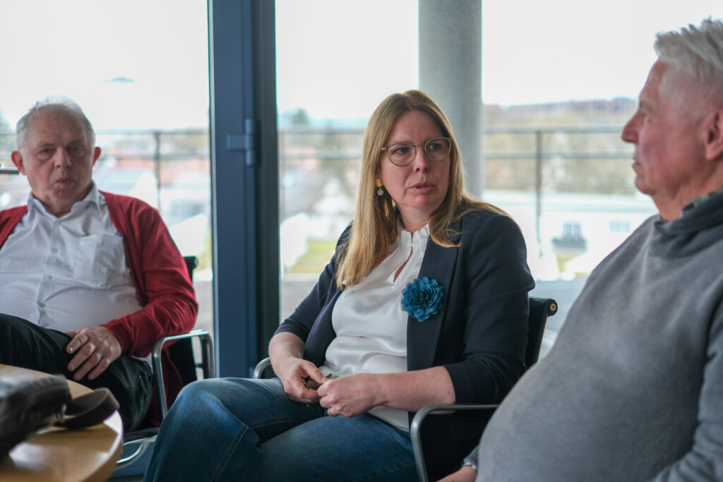 Das Bild zeigt drei Personen, die an einem runden Tisch zusammen sitzen. Eine Frau in der Mitte, links und rechts daneben jeweils ein Mann. Die Frau spricht mit dem man zu ihrer Linken und beantwortet ihm eine Frage. 