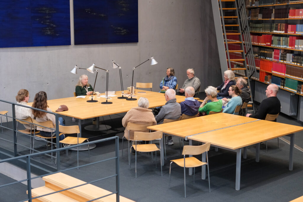 Eine Gruppe von Menschen verschiedener Altersklassen sitzt um einen Tisch im Lesesaal der deutschen Nationalbibliothek in Frankfurt am Main. In der Mitte des Tisches stehen mehrere große Leselampen. An der langen Seite des Tisches, den übrigen Menschen gegenüber, sitzt eine Frau, die aus einem Buch vorliest. Im Hintergrund sieht man eine hohe Bücherwand mit alt aussehenden Medienwerken.
