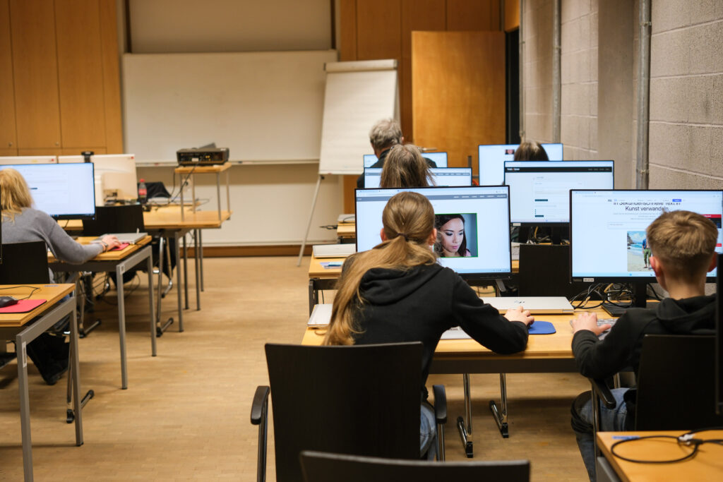 Ein geschlossener Raum, in dem zwei Reihen Tische stehen. Auf jedem der Tische stehen jeweils zwei Computer mit Monitoren. Alle sind eingeschaltet. An den Monitoren auf der rechten Seite des Vordergrundes sitzen zwei Jugendliche und bearbeiten Bilder. Sie kehren dem Betrachter den Rücken zu.